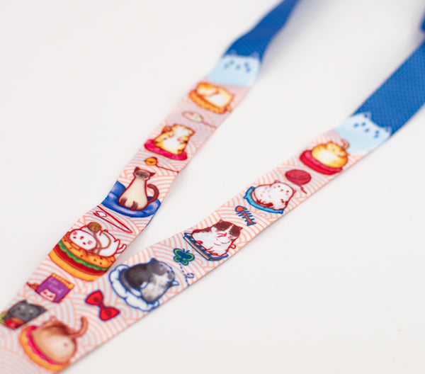 Cat lanyard with half blue dotted pattern and half light blue Japanese wave pattern. There are designs of various breeds of cats sitting and relaxing on cat cushions. For example, one tuxedo kitty is standing up and trying to catch a butterfly. A Ragdoll kitten grooming itself. A Siamese kitten being an absolute queen and sitting gracefully. A gray kitten enjoying a bowl of cat snack from its human butler. The lanyard have a lobster claw clasp to hold your badges.