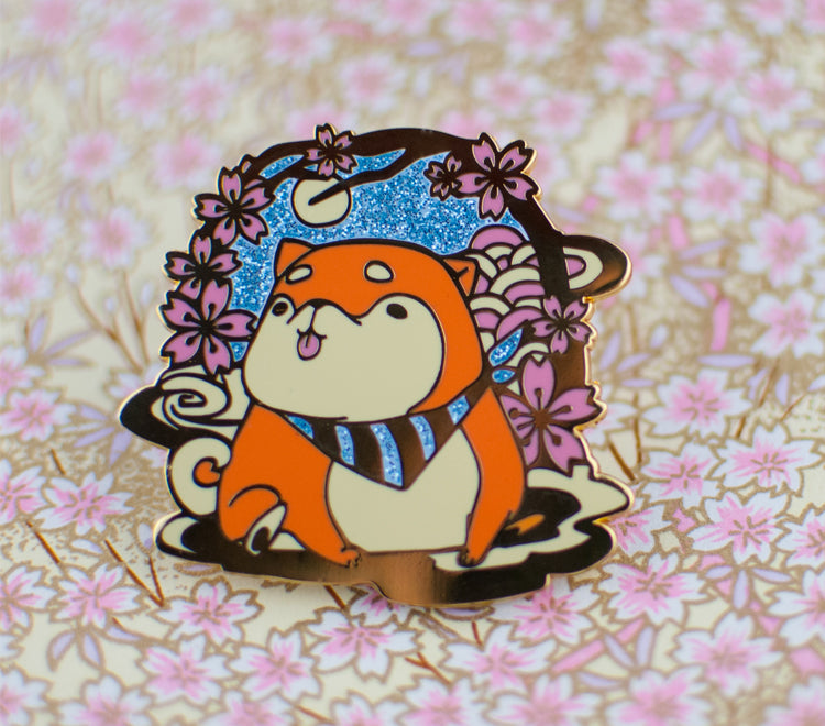 This pin features a shiba inu sitting under a sakura or cherry blossom branch enjoying the flowers under the moon. The background is a sparkly blue sky that frames the shiba inu. The pin comes in gold metal and have two rubber pin backs where you can attach the pin to other objects like your clothes or bags.